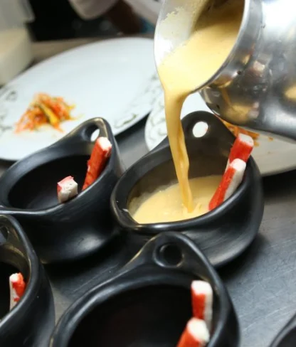 Creamy yellow soup is being poured from a metal pot into black bowls lined up in a row, each containing a breadstick.