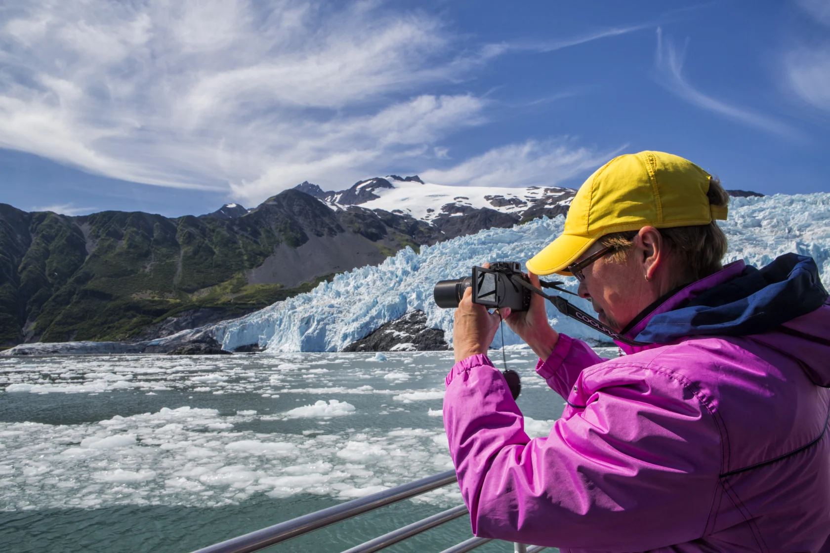 A person in a yellow cap and pink jacket takes a photo of a glacier and icy water from a boat on a sunny day.