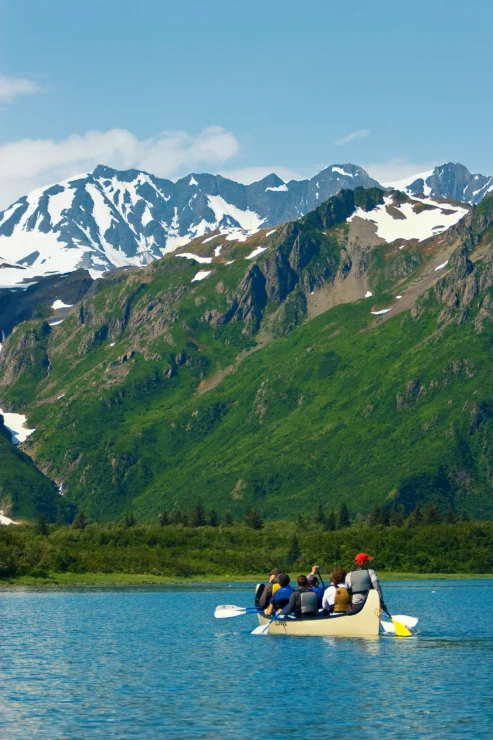 A group of people in a canoe paddles on a lake with snow-capped mountains and lush green hills in the background under a clear blue sky.