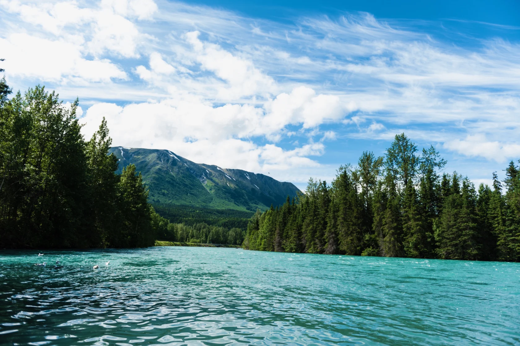 A turquoise river flows between lush green trees with mountains in the background under a cloudy blue sky.