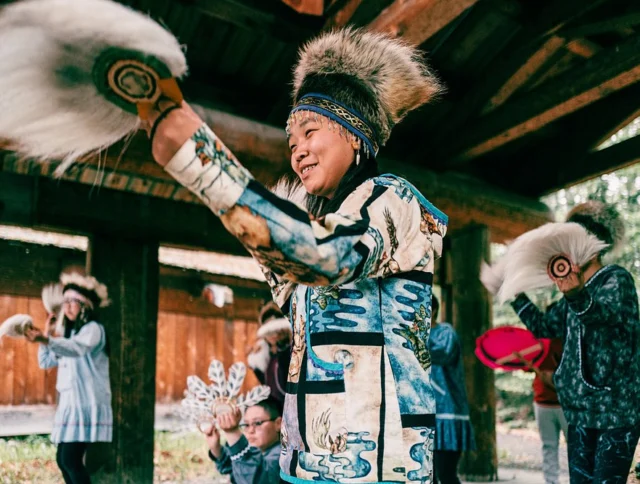 People in traditional attire perform a cultural dance under a wooden structure, holding fans and wearing fur hats.