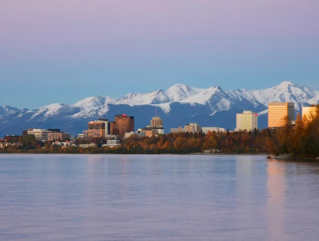 City skyline at dusk with snow-capped mountains in the background and a calm body of water in the foreground.