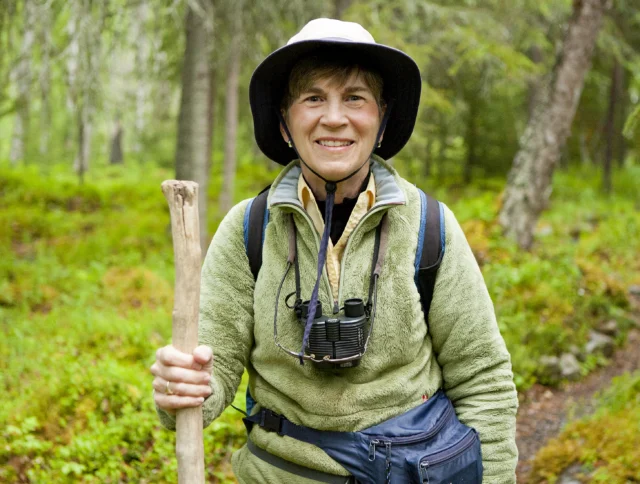 Person in a green jacket and hat hiking in a forest, holding a walking stick, with binoculars around the neck.