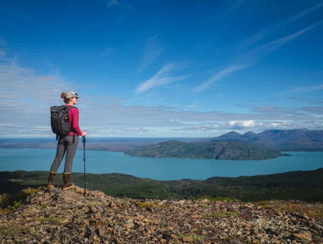 A person in hiking gear stands on a rocky hilltop, gazing at a wide view of a lake and distant mountains under a clear blue sky.