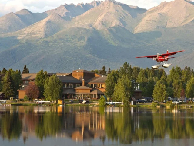 A red seaplane flies over a calm lake with reflections of trees and a large building. Mountains are visible in the background under a partly cloudy sky.