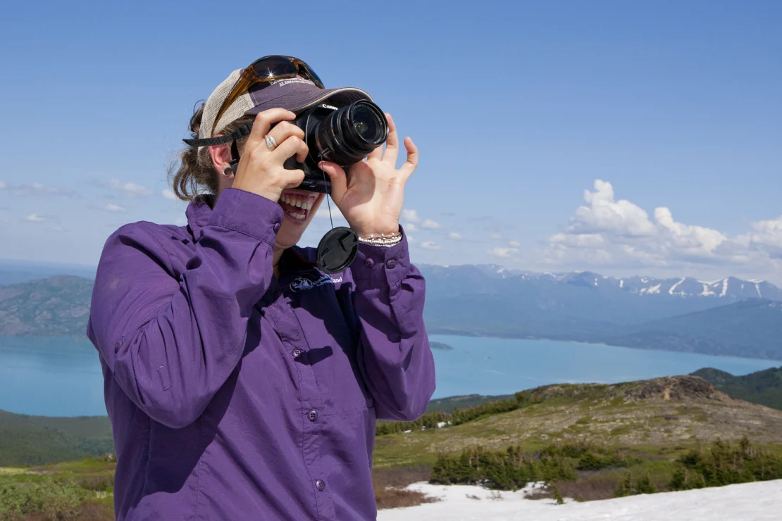 Person in a purple jacket takes a photo with a camera on a mountain, overlooking a lake and distant snow-capped peaks under a clear blue sky.