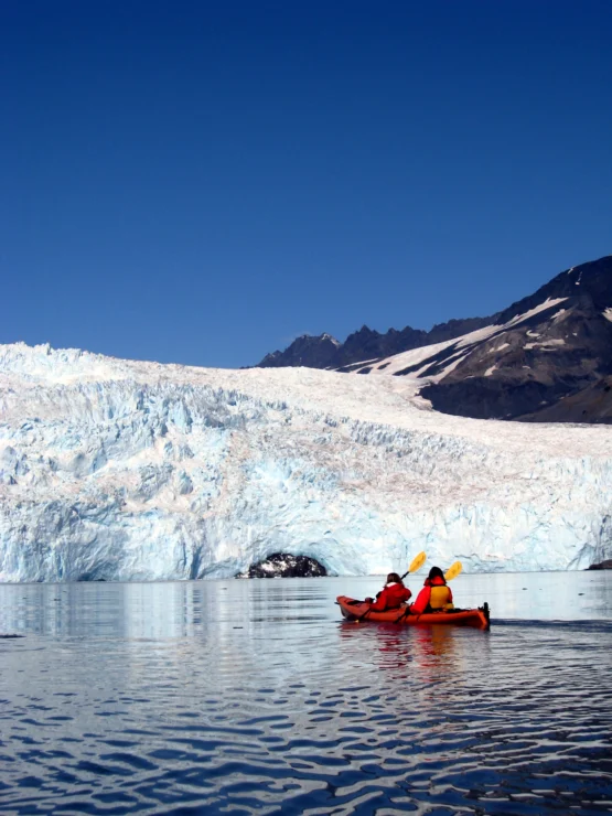 Two people in red kayaks paddle on calm water near a large glacier under a clear blue sky.