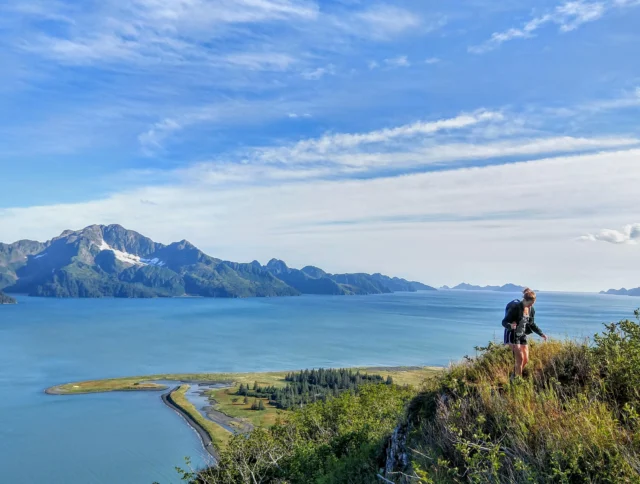 Person with a backpack stands on a hilltop, overlooking a scenic coastal landscape with mountains and a blue sky in the background.