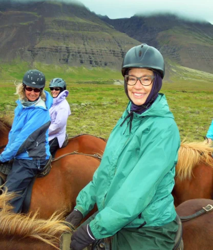 Four people riding horses in a grassy landscape with mountains in the background, all wearing helmets and outdoor jackets.