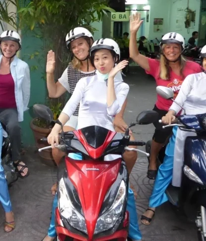 A group of people wearing helmets on scooters, smiling and waving at the camera.