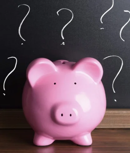 A pink piggy bank sits on a wooden surface in front of a blackboard with drawn question marks.