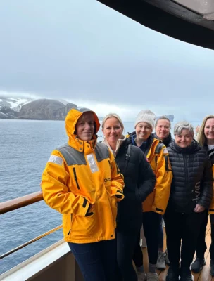 A group of people in yellow jackets stands on a ship's deck with a snowy landscape and ocean in the background.