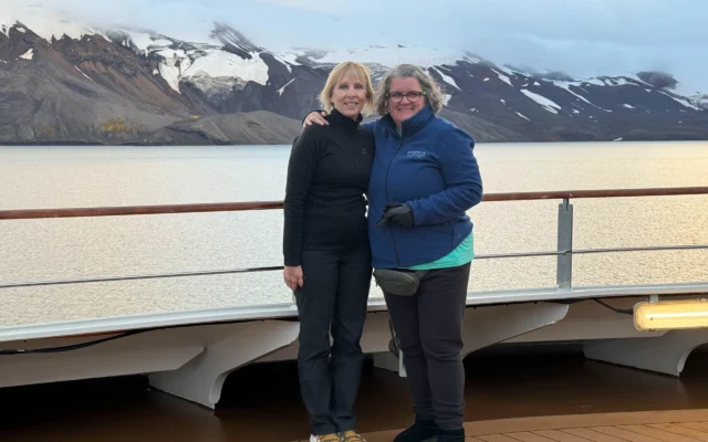 Two women stand on a boat deck with a scenic backdrop of mountains and water.