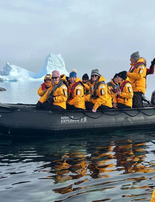 A group of people wearing yellow jackets sit in an inflatable boat on calm water with icebergs in the background.