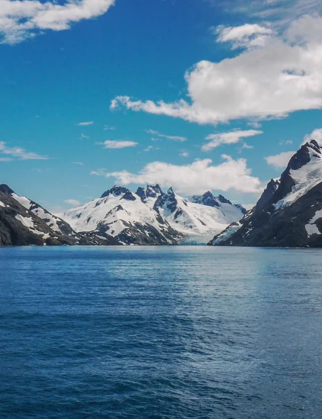 A view of snow-capped mountains and calm blue water under a partly cloudy sky.