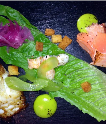 A gourmet dish with smoked salmon, lettuce, pickled onions, fish filet, green sauce, and croutons, arranged on a black plate.