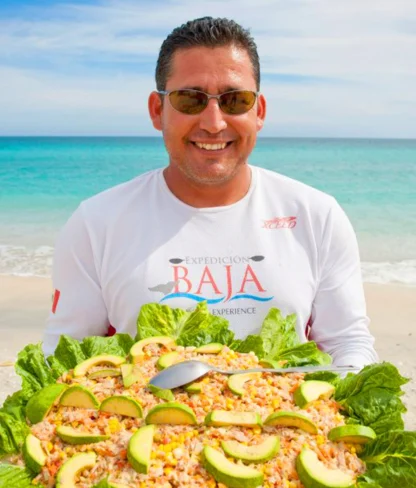 A man in sunglasses stands on a beach, holding a large platter of food topped with avocado slices and lettuce.