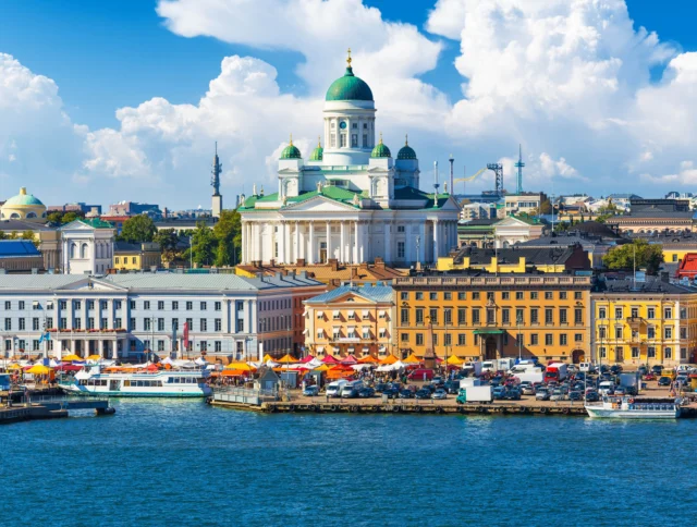 Scenic view of Helsinki with Helsinki Cathedral in the background, colorful market stalls, and boats docked by the waterfront under a partly cloudy sky.