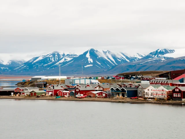 Coastal village with colorful houses near a calm body of water, set against a backdrop of snow-capped mountains and a cloudy sky.