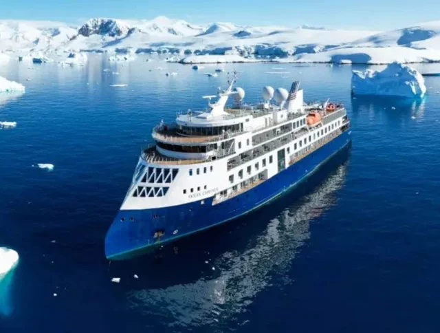 A blue and white cruise ship navigates through icy waters surrounded by icebergs under a clear blue sky.