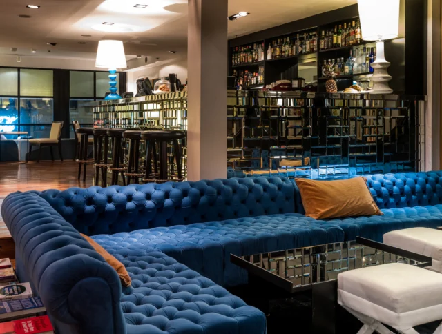 Stylish lounge with a blue tufted sofa, mirrored tables, and tall bar area in the background. Warm lighting creates a cozy atmosphere.