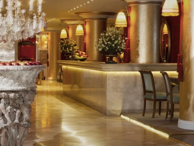 Opulent hotel lobby with marble floors, ornate columns, a decorative stone pedestal with apples, elegant chairs, a chandelier, and floral arrangements on the reception desk.