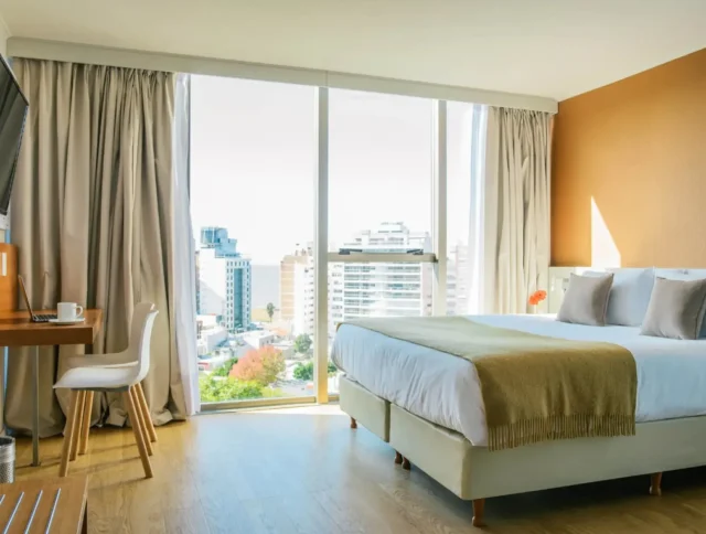 Modern hotel room with a large bed, a desk with a chair, and a wall-mounted TV. Floor-to-ceiling windows offer a city view, while curtains frame the scene. A light brown accent wall adds warmth.