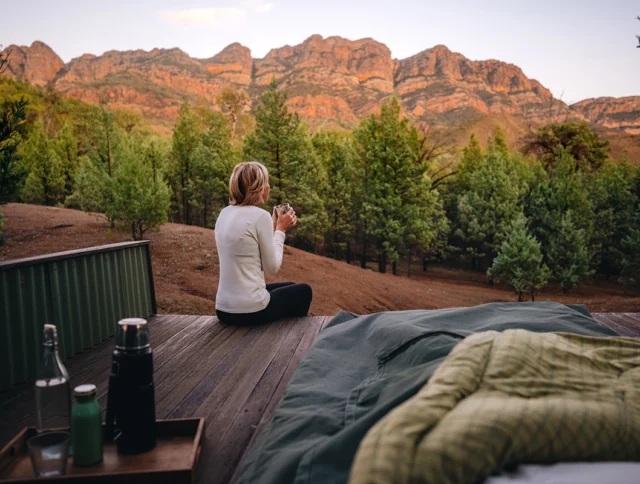 A person sits on a wooden deck, sipping a drink and overlooking a forest with mountains in the background. A bed and a tray with bottles are nearby.