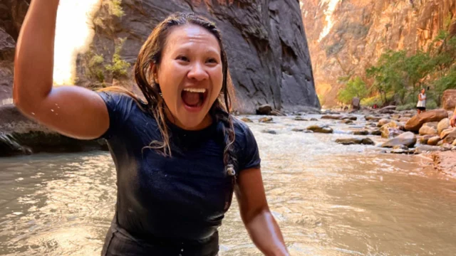 A woman in a wet black shirt and white shorts raises her fist in excitement while standing in a rocky river canyon.