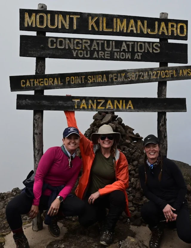 Three people pose in front of a Mount Kilimanjaro sign indicating they are at Cathedral Point Shira Peak, Tanzania.