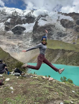 Person jumping joyfully in front of a turquoise lake and snow-capped mountains, while others sit nearby.