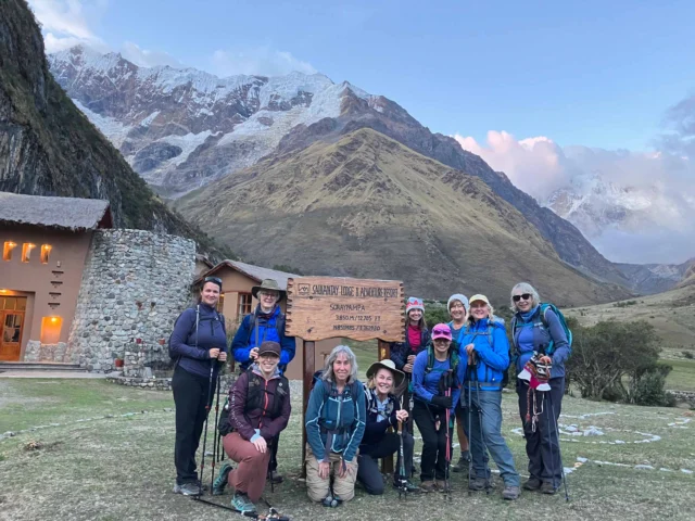 Group of hikers with trekking poles posing in front of a sign in a mountainous landscape with snow-capped peaks in the background.
