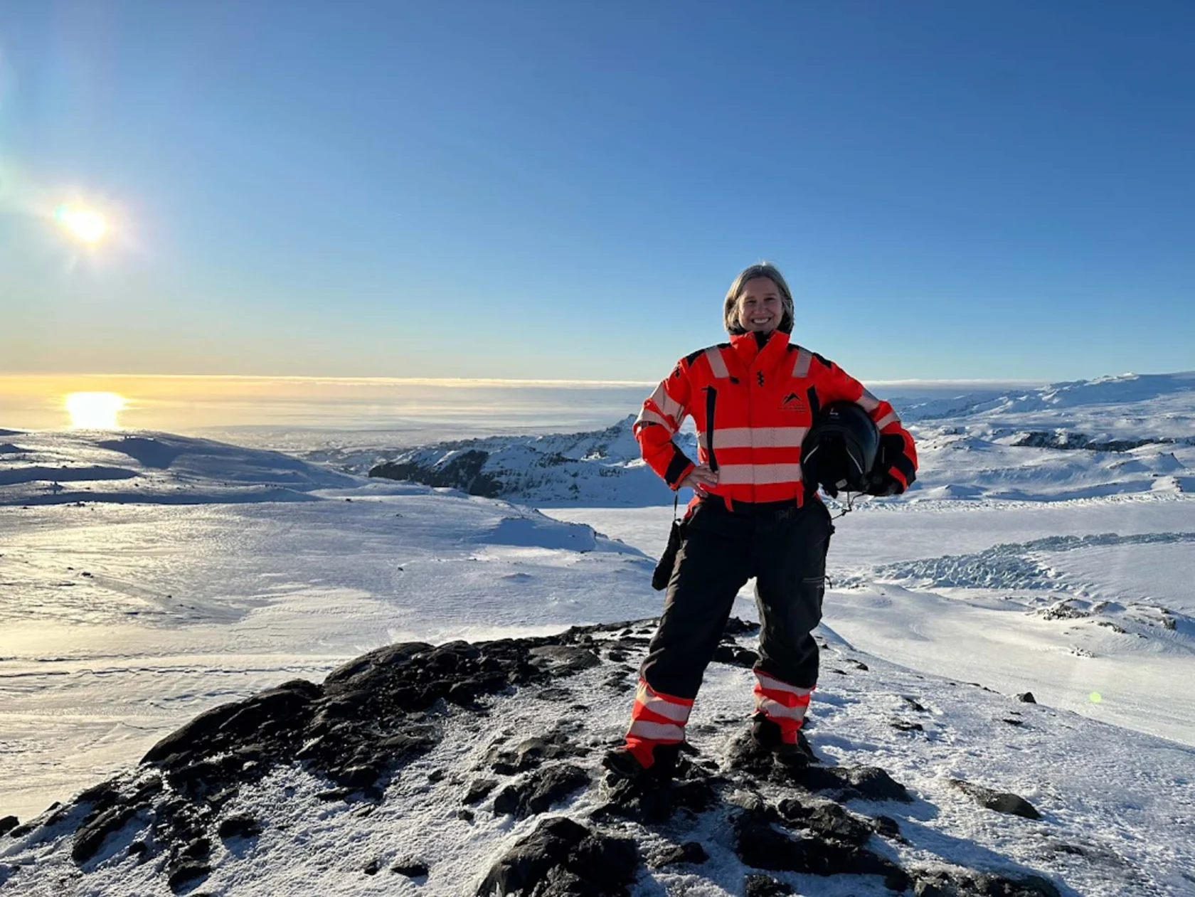 A person in red and black winter gear stands on a snowy hill with a clear blue sky and distant sunset.