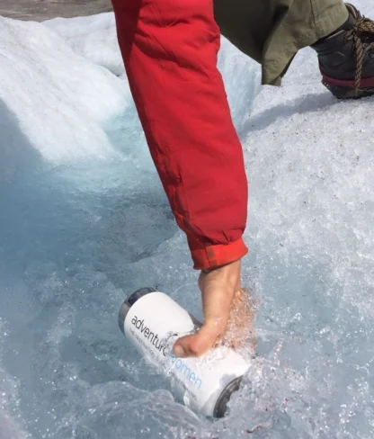 A person in a red jacket and hiking boots places a white water bottle in a stream on icy terrain.