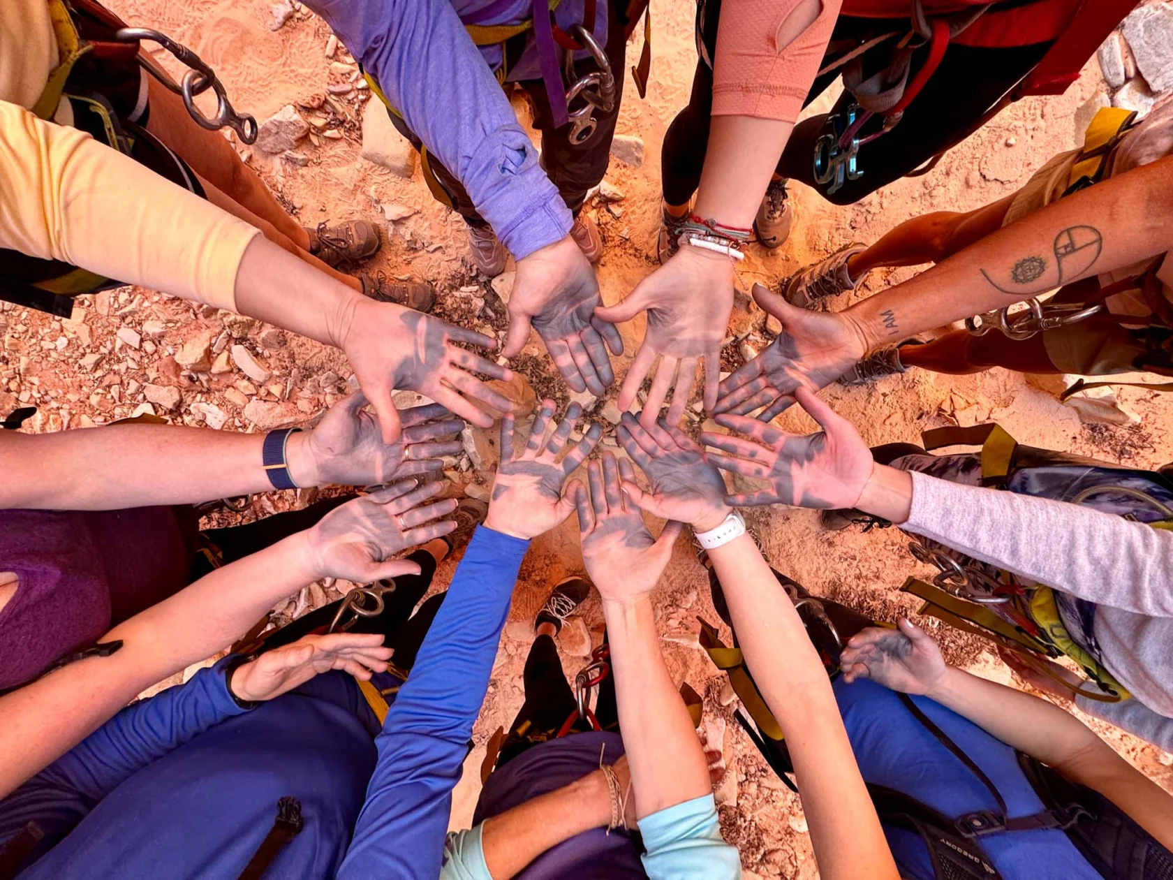 A group of people standing in a circle, with their hands extended toward the center, showing dirt-covered palms. They're outdoors on reddish rocky ground.