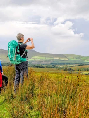 Two hikers with backpacks stand on a grassy hill, looking at a landscape view of rolling hills and fields under a partly cloudy sky.