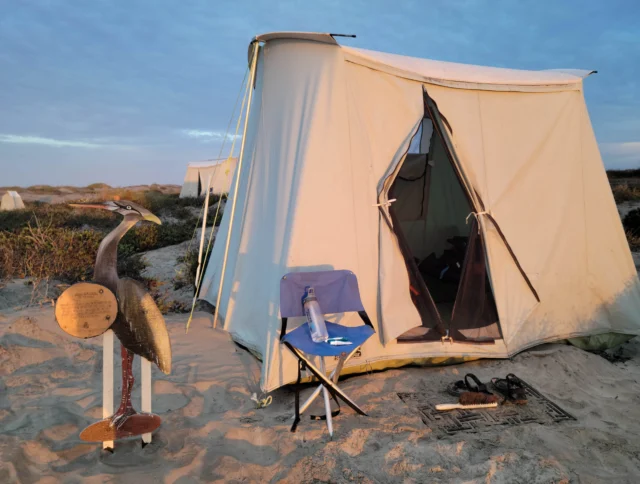 A beige tent with an open entrance stands on sandy ground. In front, there's a chair with a water bottle and shoes on the ground. A bird sculpture is on the left. The sky is partly cloudy.