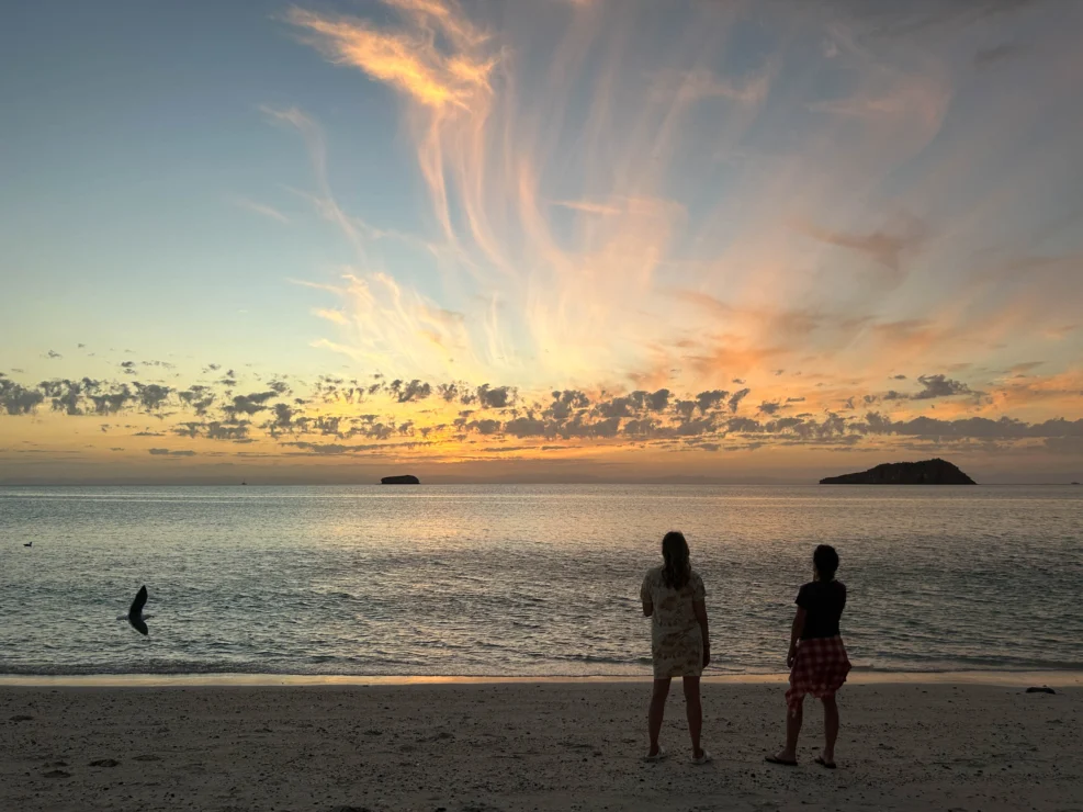 Two people stand on a beach at sunset, facing the ocean. The sky displays wispy clouds with orange and blue hues. Small islands are visible in the distance.