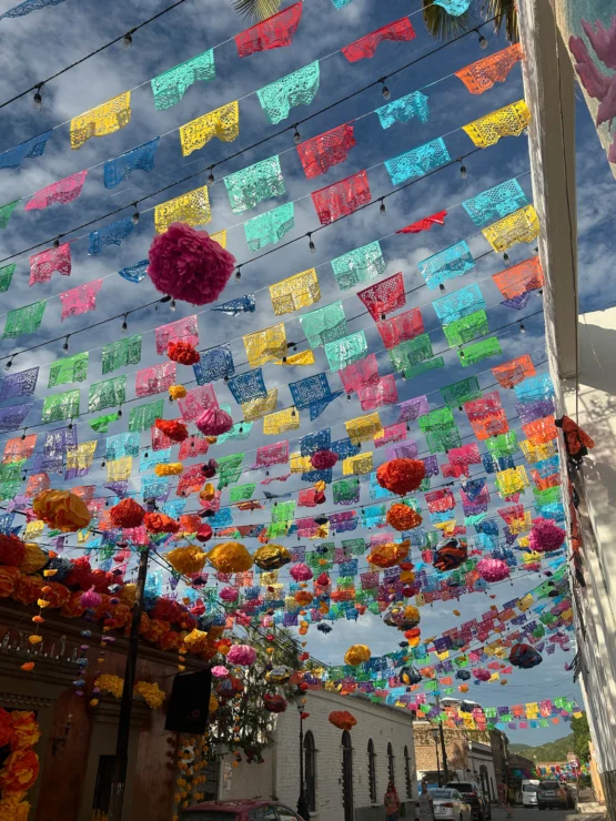 Colorful papel picado and pom-pom decorations hang above a street, with blue sky visible. Buildings line the street below.