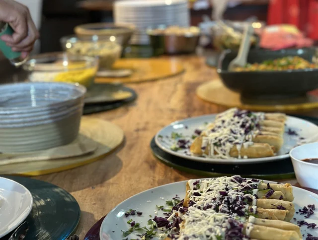 A table is set with plates of rolled tortillas topped with cheese and garnishes. Bowls of various sides and dishes are in the background. A person reaches for a serving.