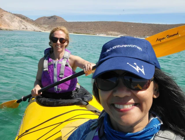 Two people kayaking on clear turquoise water near rocky shores. The foreground person wears a cap and sunglasses; both are in purple life jackets. The sky is clear.