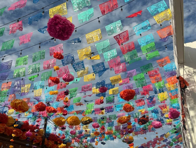 Colorful papel picado and flower decorations hang across a street under a blue sky.