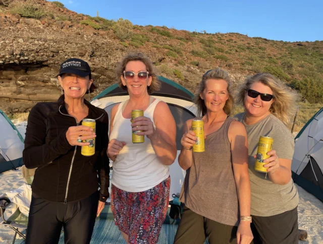 Four women stand in front of tents on a beach, holding canned drinks and smiling. The background shows sandy terrain and vegetation.