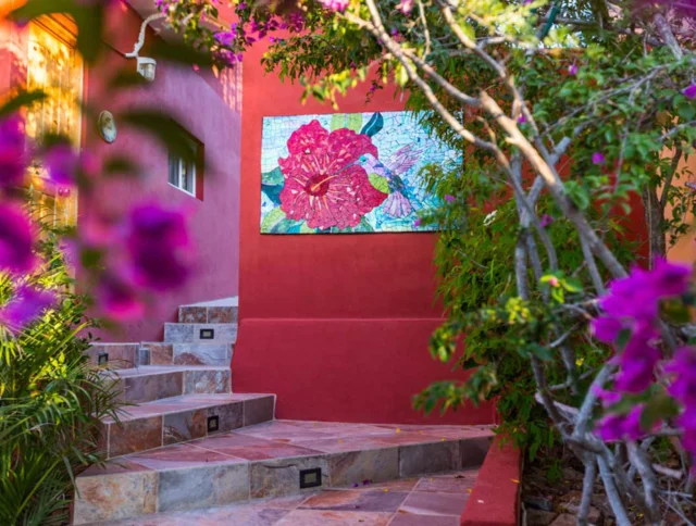 Outdoor stairway with colorful tiled steps leads to a red wall adorned with a large floral mosaic. Purple flowers frame the scene.