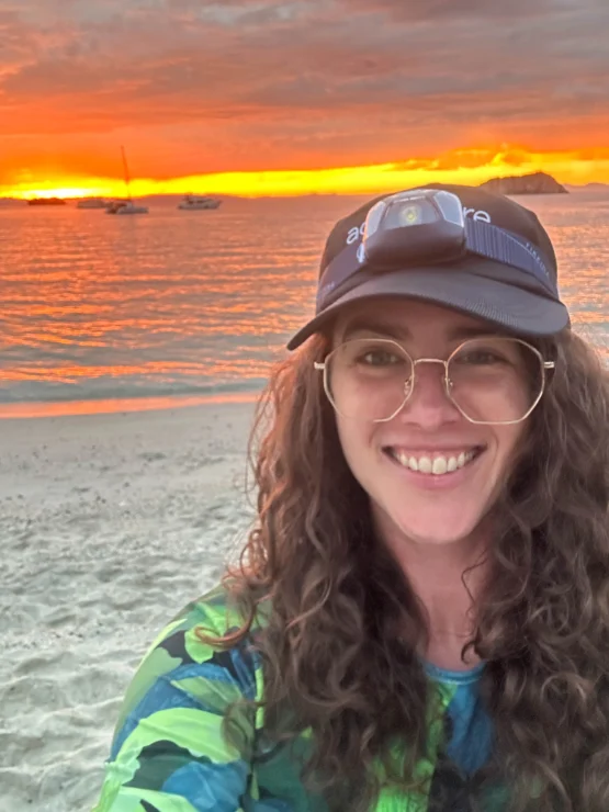 A person with curly hair and glasses, wearing a cap with a headlamp, smiles on a beach at sunset. Boats are visible on the water.