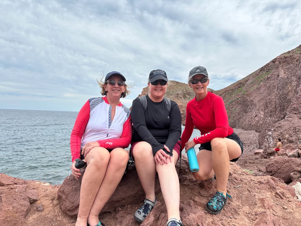 Three people sit on rocks by the sea, wearing casual outdoor clothing with hats and sunglasses. Rocky hills are in the background under a cloudy sky.