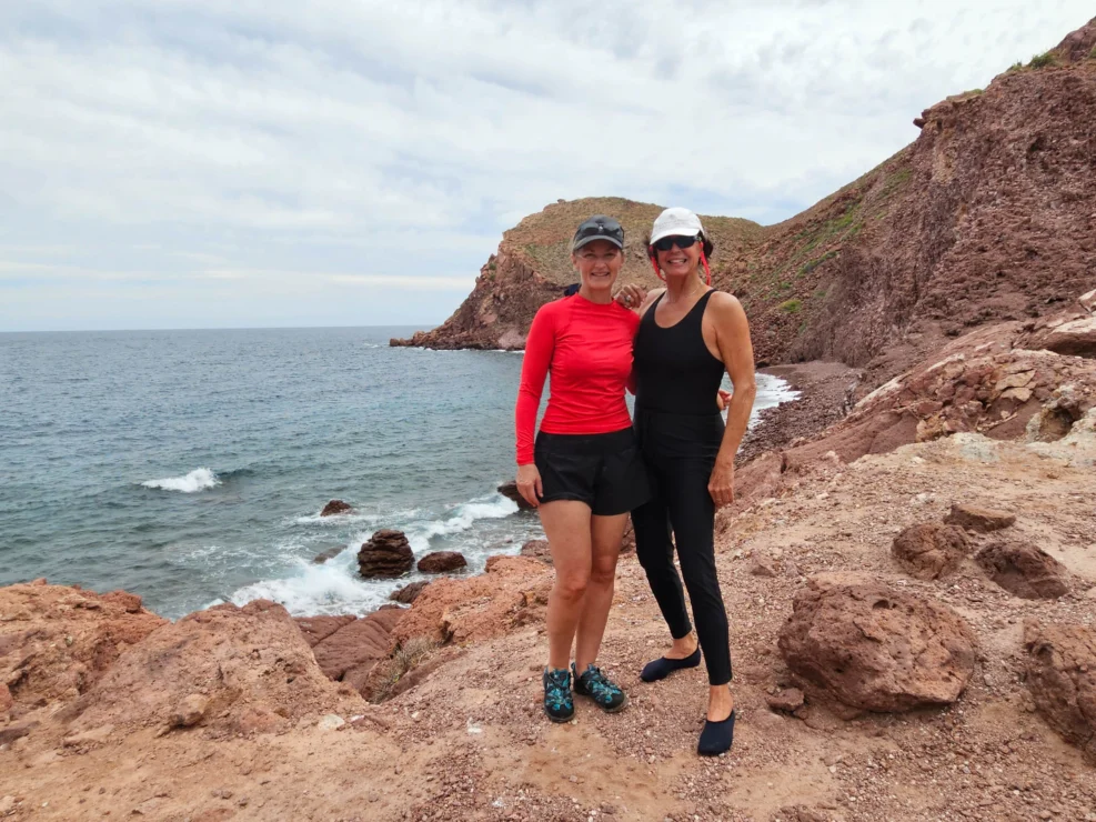 Two women in athletic wear stand on rocky terrain by the ocean, with cliffs in the background.