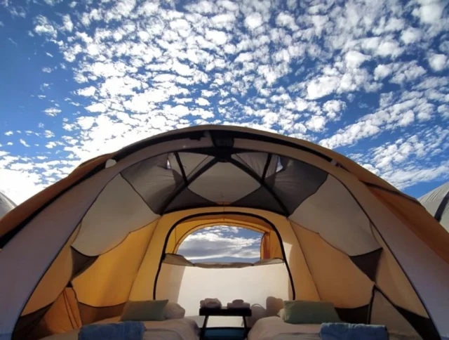Interior view of a tent with two beds and a small table, set under a sky with scattered clouds.