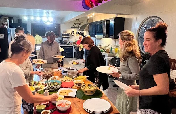 A group of people serving themselves food from a buffet-style table in a kitchen with colorful caps on a shelf above.