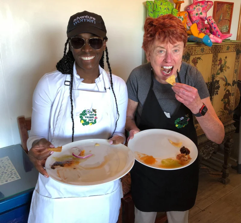 Two women enjoying food; one holds an empty plate with a chip, the other eats and holds another empty plate. They are standing indoors and wearing aprons.